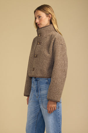 A person stands against a beige background wearing the ESME FAUX SHERPA REVERSIBLE JACKET by Velvet by Graham & Spencer—a cropped, high-neck brown wool jacket with large buttons and luxe faux suede detailing that adds texture and warmth—paired with blue high-waisted jeans.