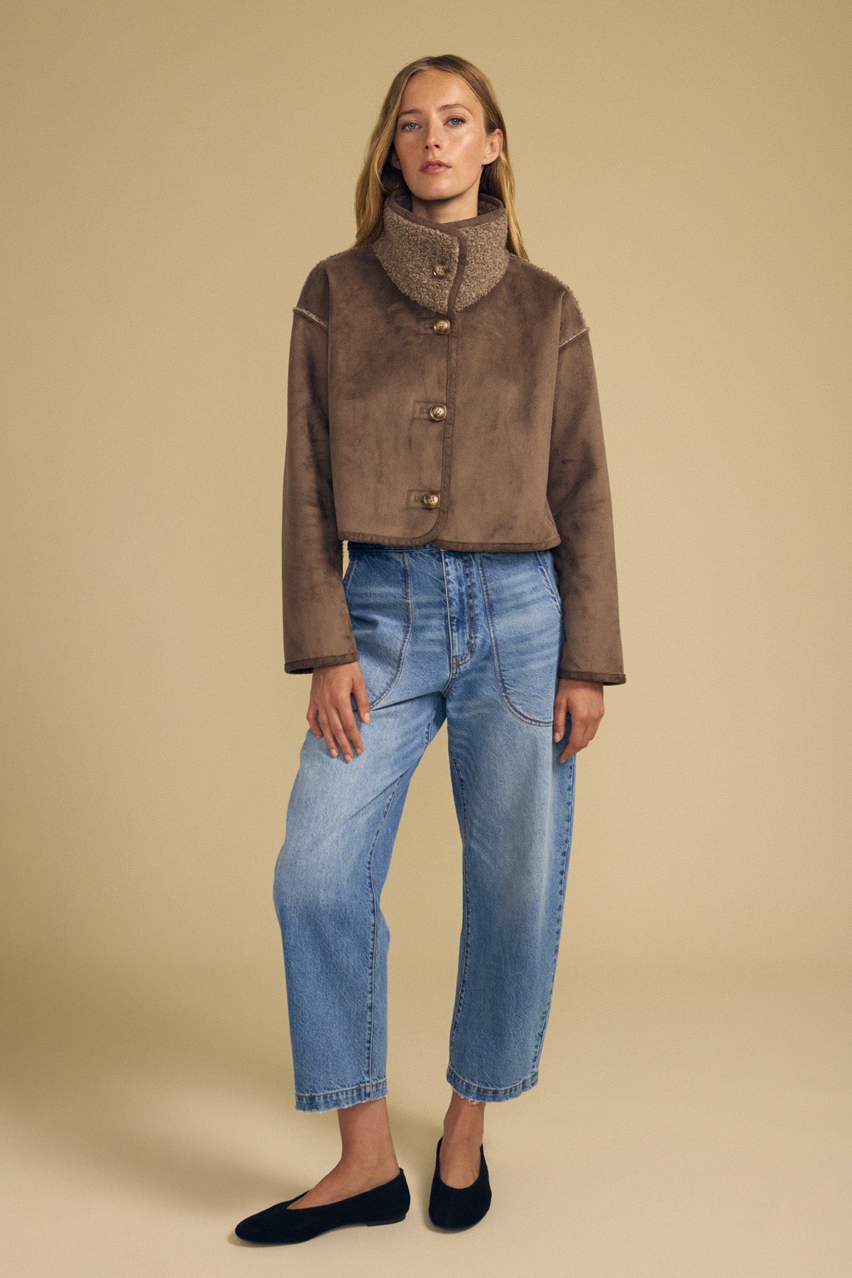 A woman stands against a beige background wearing the ESME FAUX SHERPA REVERSIBLE JACKET by Velvet by Graham & Spencer, paired with loose blue jeans and black flats.