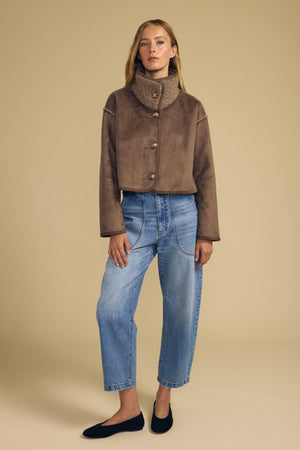 A woman stands against a beige background wearing the ESME FAUX SHERPA REVERSIBLE JACKET by Velvet by Graham & Spencer, paired with loose blue jeans and black flats.