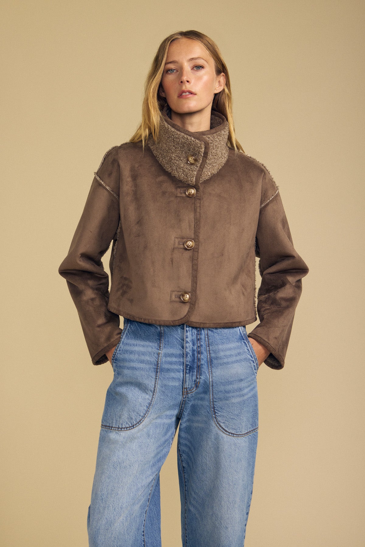 A woman stands with her hands in her pockets against a beige background, wearing the Velvet by Graham & Spencer ESME Faux Sherpa Reversible Jacket in brown, featuring a high collar and buttoned front, paired with light blue jeans.