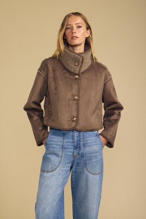 A woman stands with her hands in her pockets against a beige background, wearing the Velvet by Graham & Spencer ESME Faux Sherpa Reversible Jacket in brown, featuring a high collar and buttoned front, paired with light blue jeans.