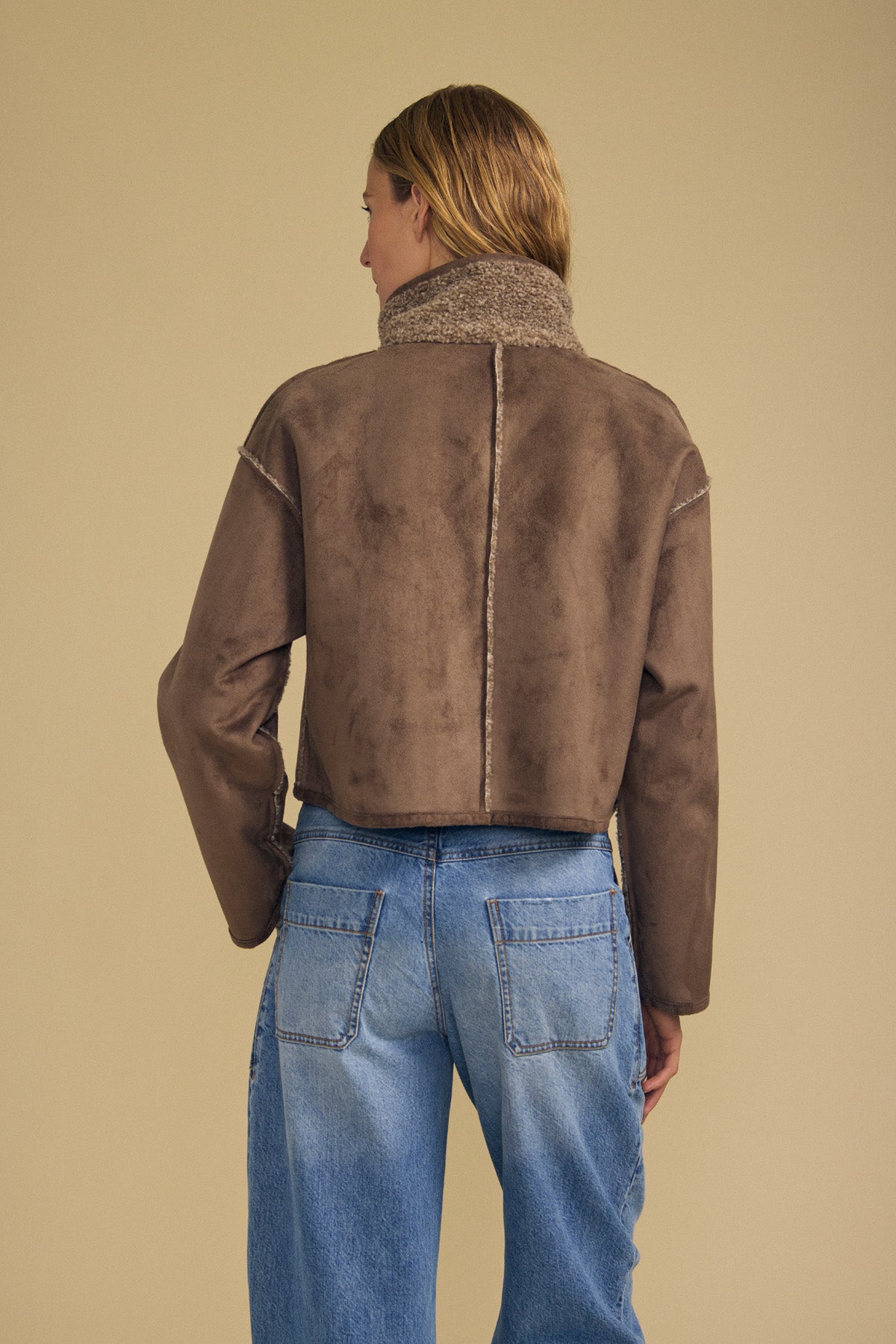 A person with light brown hair, seen from behind, wears the ESME FAUX SHERPA REVERSIBLE JACKET by Velvet by Graham & Spencer and blue jeans, standing against a beige background.