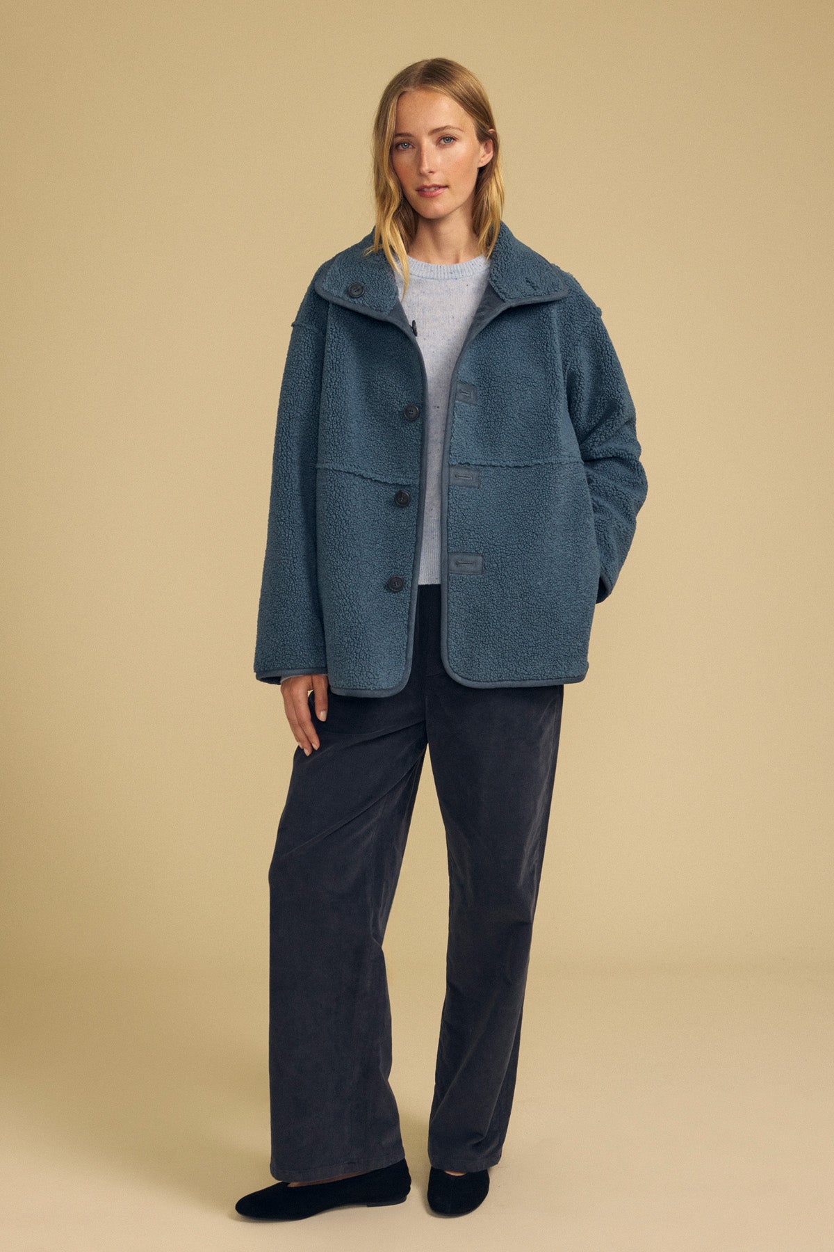 A woman stands against a beige background wearing the SHONA FAUX SHERPA REVERSIBLE JACKET by Velvet by Graham & Spencer layered over a light sweater, paired with dark wide-leg pants and black shoes.-41964712231105