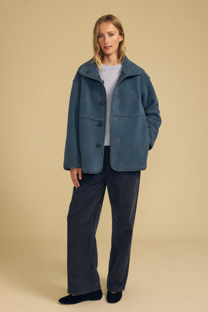 A woman stands against a beige background wearing the SHONA FAUX SHERPA REVERSIBLE JACKET by Velvet by Graham & Spencer layered over a light sweater, paired with dark wide-leg pants and black shoes.