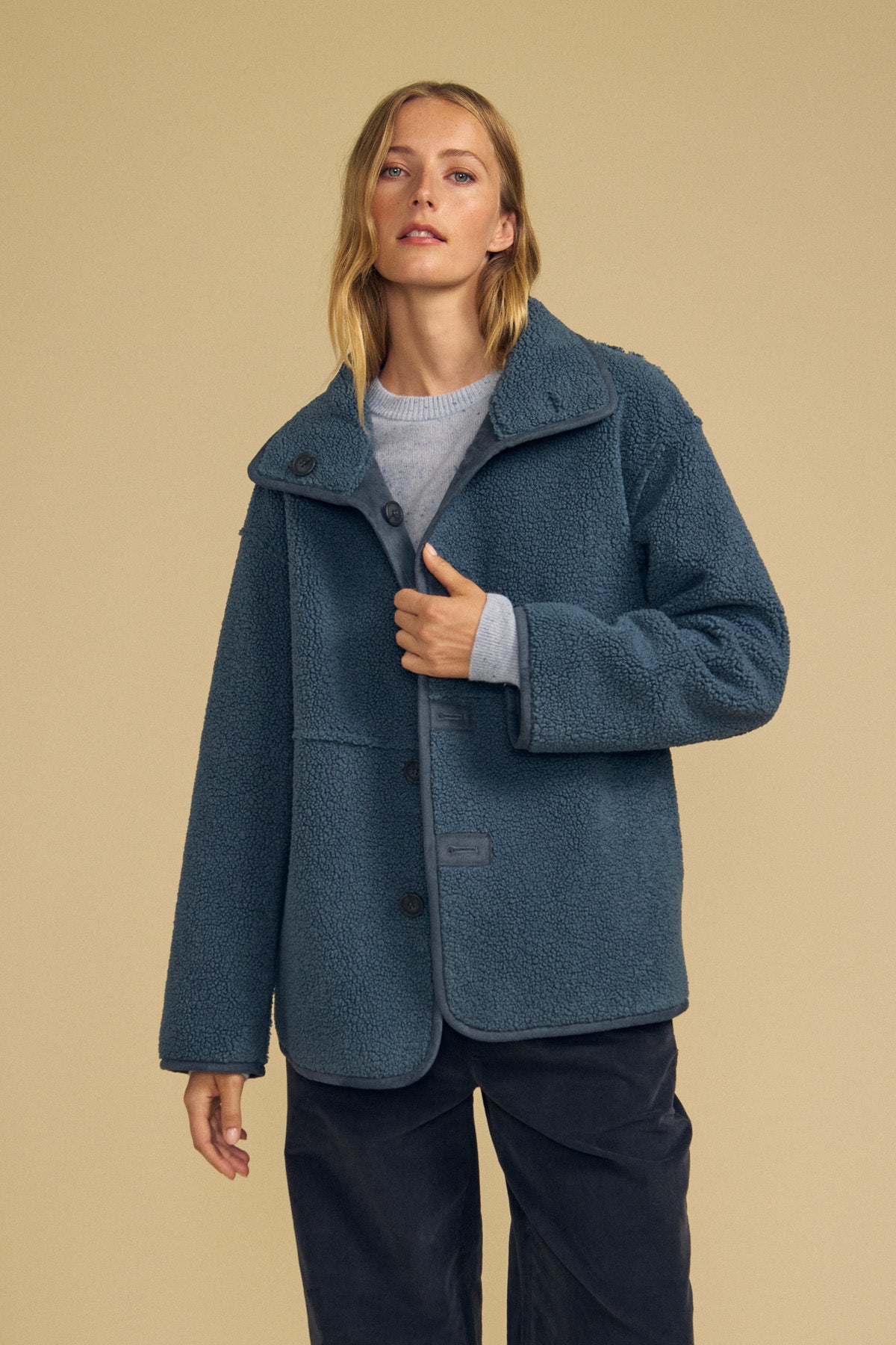 A person stands against a plain beige background wearing the Velvet by Graham & Spencer SHONA Faux Sherpa Reversible Jacket in blue, paired with a gray top and dark pants.