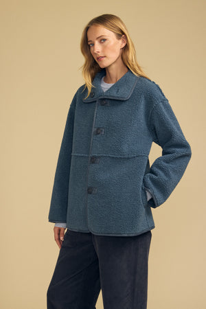 A woman stands against a plain beige background wearing the SHONA FAUX SHERPA REVERSIBLE JACKET by Velvet by Graham & Spencer—a blue, textured button-up style with wide sleeves—paired with dark pants.