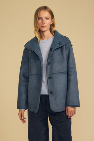Wearing the SHONA FAUX SHERPA REVERSIBLE JACKET by Velvet by Graham & Spencer, a person stands against a plain beige background. The blue faux suede jacket features button closures and a textured sherpa collar, layered stylishly over a light sweater.