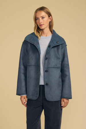 A woman wearing the SHONA FAUX SHERPA REVERSIBLE JACKET by Velvet by Graham & Spencer in blue, layered over a light gray sweater and dark pants, stands against a beige background.