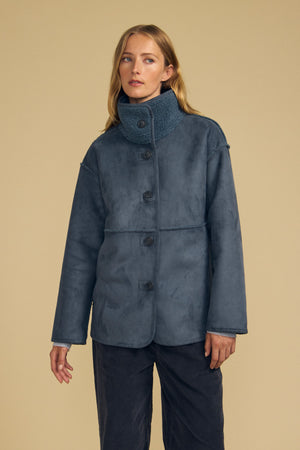 A woman wearing the Velvet by Graham & Spencer SHONA FAUX SHERPA REVERSIBLE JACKET in blue, styled with dark blue pants, stands against a beige background.