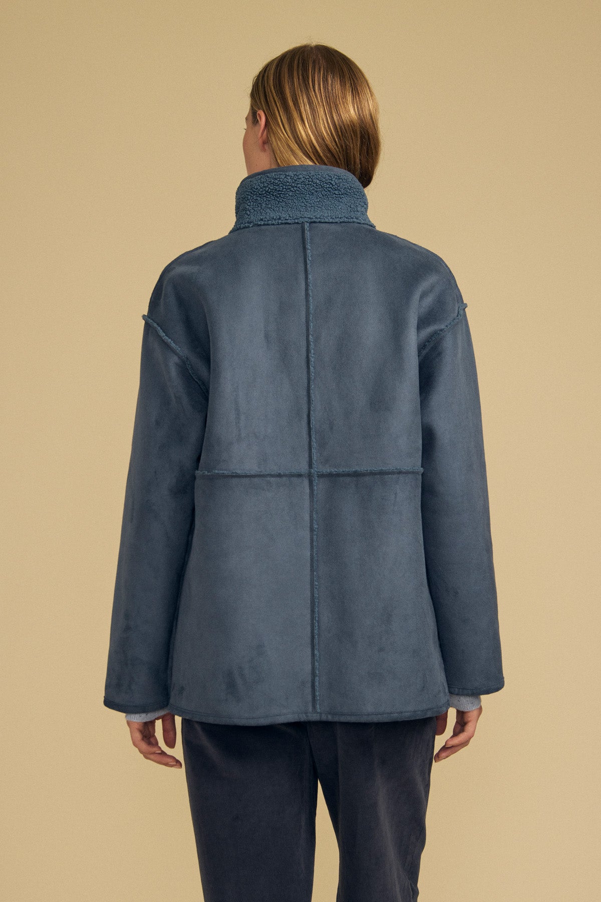 A person with straight light brown hair is shown from the back, standing against a beige background, wearing the SHONA FAUX SHERPA REVERSIBLE JACKET by Velvet by Graham & Spencer in blue faux suede shearling, paired with matching pants.