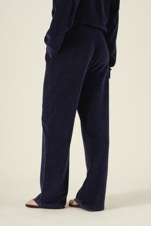 A person viewed from behind wearing navy blue DAISY VELOUR TRACK PANT lounge pants with a plush feel and flip-flops.