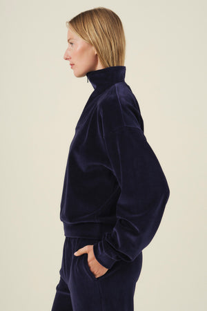 Profile view of a person wearing a navy blue DANICA VELOUR HALF-ZIP SWEATSHIRT with a high collar and hands in their pockets.