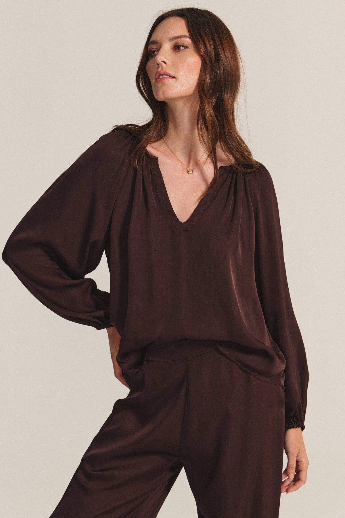   A woman wears the LYLA TOP by Velvet by Graham & Spencer—a loose, long-sleeve satin top with a deep V-neck—paired with matching brown pants, standing against a neutral background. 