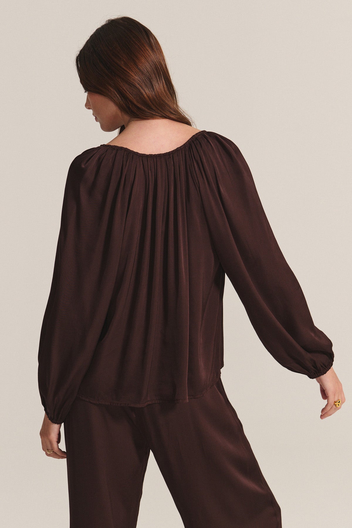 A woman faces away, wearing the LYLA TOP by Velvet by Graham & Spencer—a loose-fitting, long-sleeved dark brown satin top with subtle pleats—paired with matching pants against a plain light background.-40248149835969