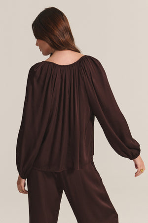 A woman faces away, wearing the LYLA TOP by Velvet by Graham & Spencer—a loose-fitting, long-sleeved dark brown satin top with subtle pleats—paired with matching pants against a plain light background.