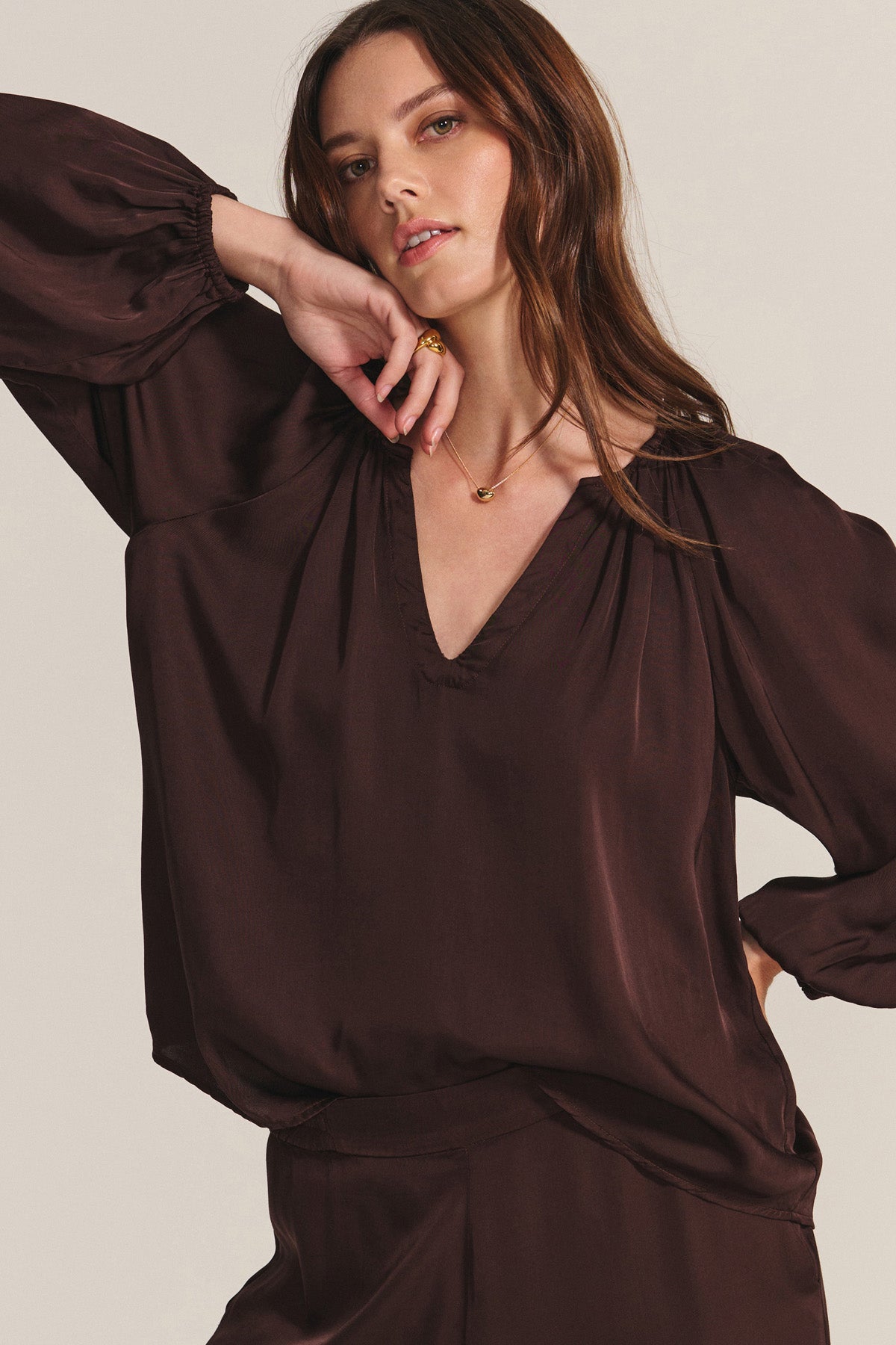A woman poses against a plain background wearing the LYLA TOP by Velvet by Graham & Spencer, a loose, long-sleeved deep brown satin blouse with pleated details and matching pants.-40248149770433