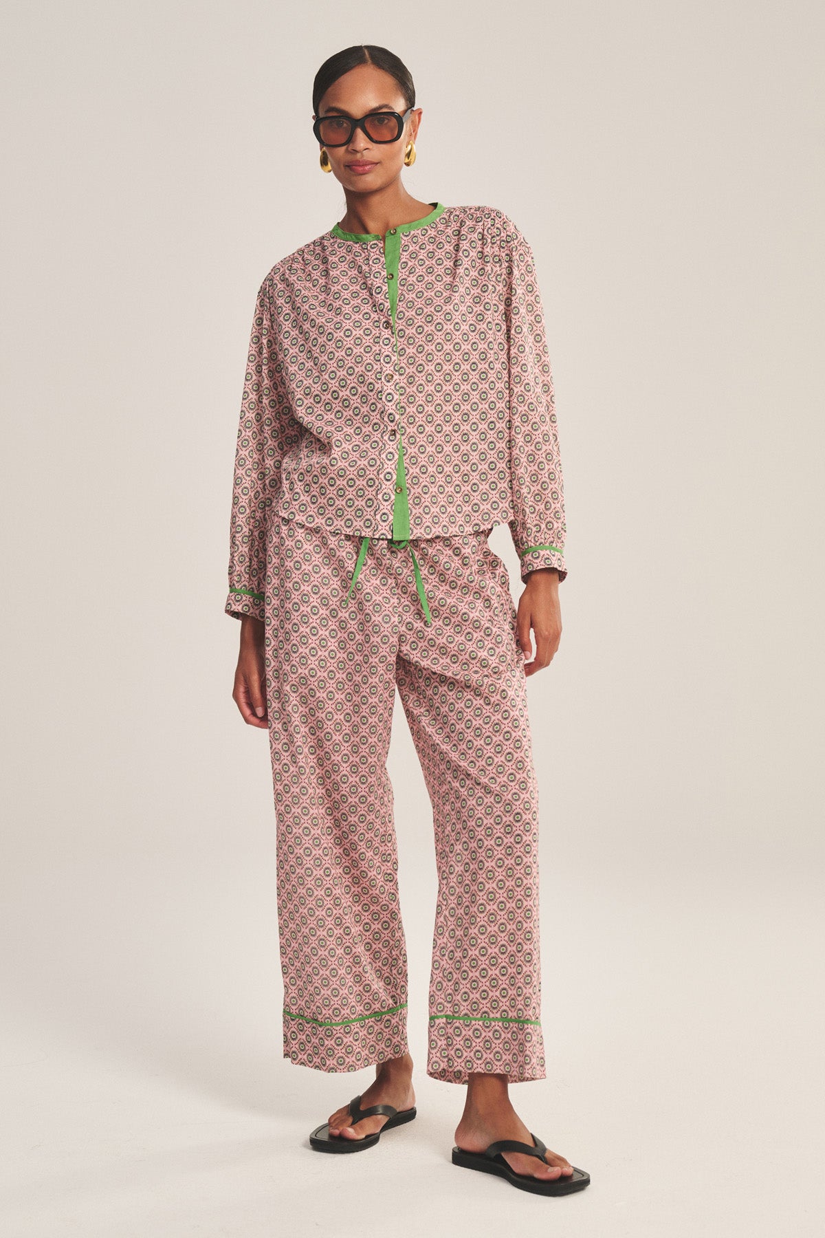 A person stands against a plain background wearing Velvet by Graham & Spencer's MABEL PANT, a patterned pink and green pajama with contrast piping, paired with black sandals, hoop earrings, and black-framed glasses.-41088992084161