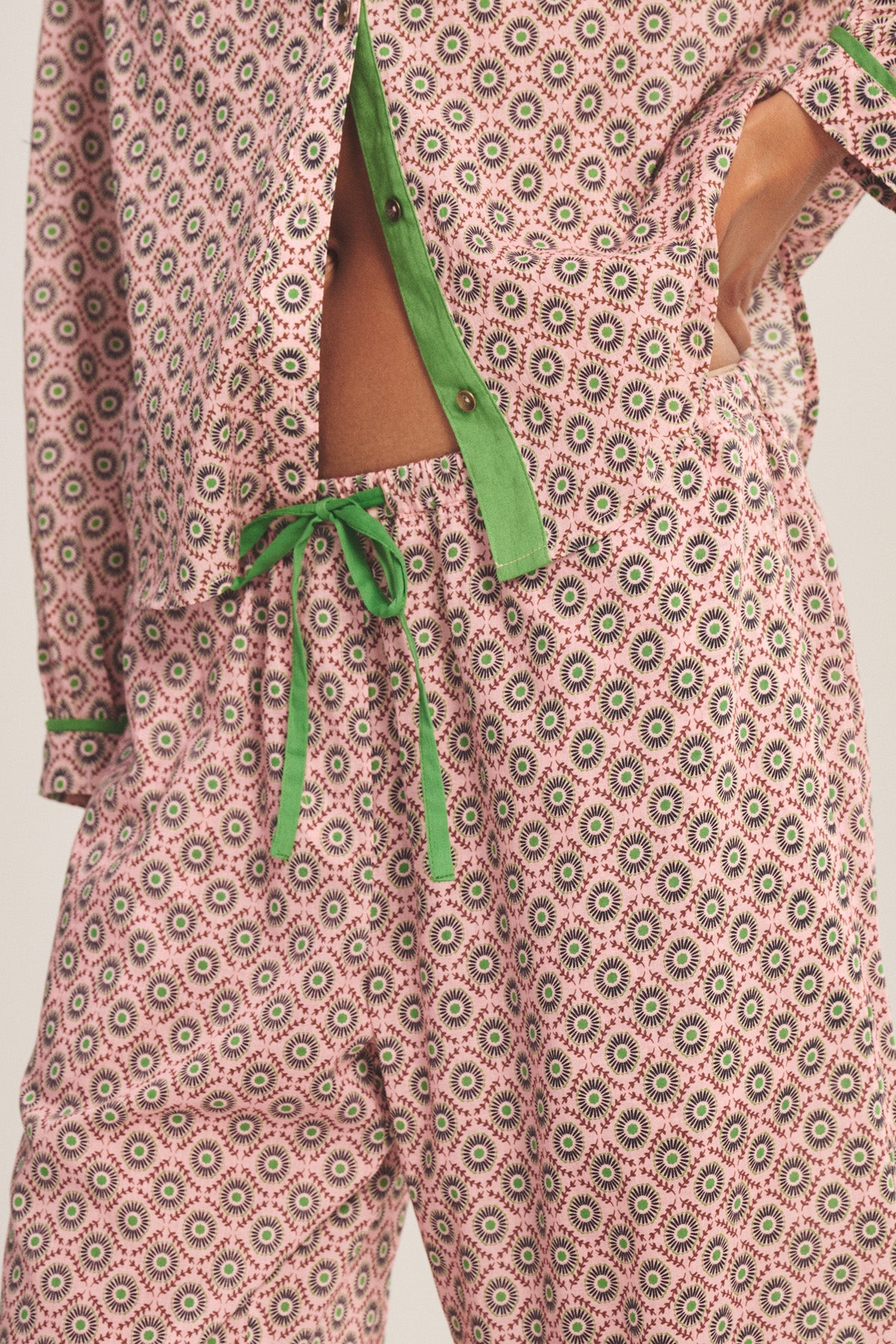 Close-up of a person wearing the Velvet by Graham & Spencer MABEL PANT—patterned pajamas with contrast piping and a green drawstring waist—showing part of their midriff with the shirt partly unbuttoned.