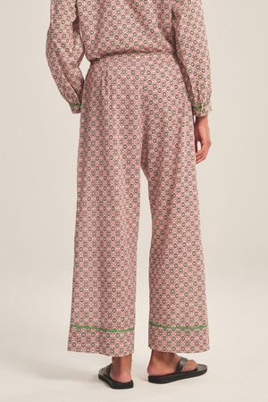 A person stands barefoot on a light background wearing the Velvet by Graham & Spencer MABEL PANT, featuring a patterned pink and white wide-leg design with a drawstring waist, paired with a matching long-sleeve top with contrast piping.