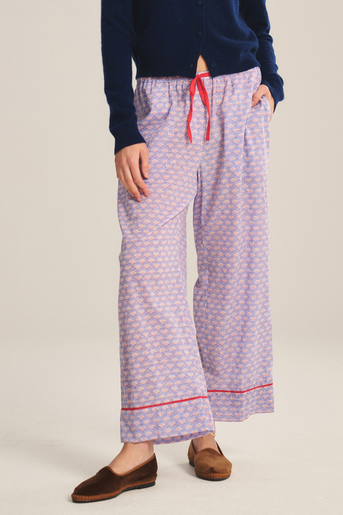 A person wearing a navy blue cardigan, Velvet by Graham & Spencer MABEL PANT with a relaxed leg and red drawstring waist, and brown slippers stands against a plain background.
