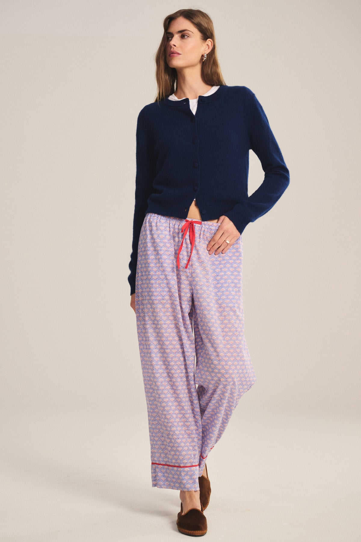 A woman stands against a plain background wearing Velvet by Graham & Spencer’s MABEL PANT, a lightweight cotton pant with a relaxed leg and red drawstring waist, paired with a navy cardigan, white shirt, and brown slippers.