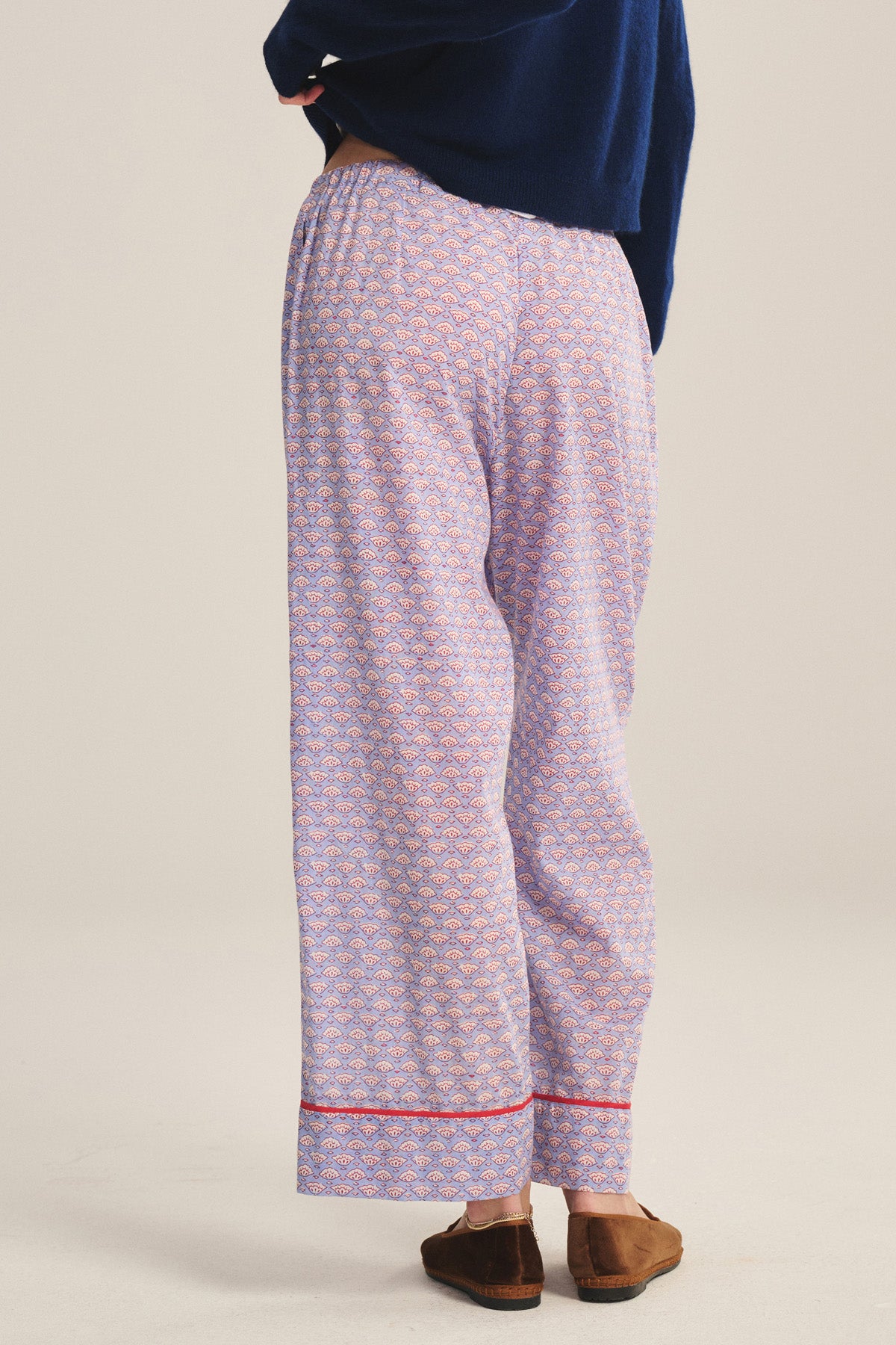 A person is shown from the back wearing the Velvet by Graham & Spencer MABEL PANT—loose, lightweight cotton pajama pants with relaxed legs, a red hem trim, and drawstring waist—paired with brown slippers and a navy blue top.