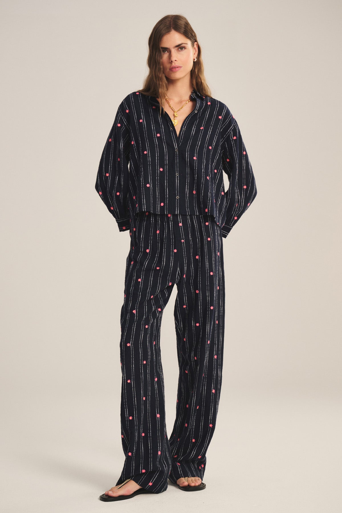A woman wears the MADELYN PANT by Velvet by Graham & Spencer—relaxed fit, dark blue with vertical white stripes and red dots—paired with a matching button-up shirt and black sandals.