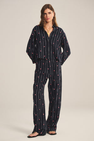 A woman wears the MADELYN PANT by Velvet by Graham & Spencer—relaxed fit, dark blue with vertical white stripes and red dots—paired with a matching button-up shirt and black sandals.