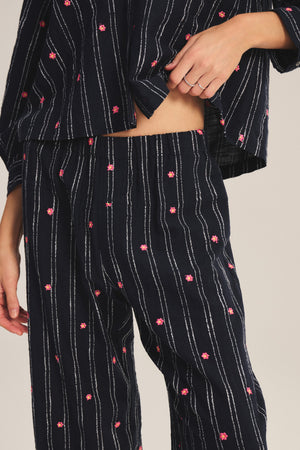 A person models the MADELYN PANT by Velvet by Graham & Spencer—a relaxed-fit, navy blue pajama pant with white vertical stripes and small pink floral patterns, paired with a matching top that reveals part of their midriff.