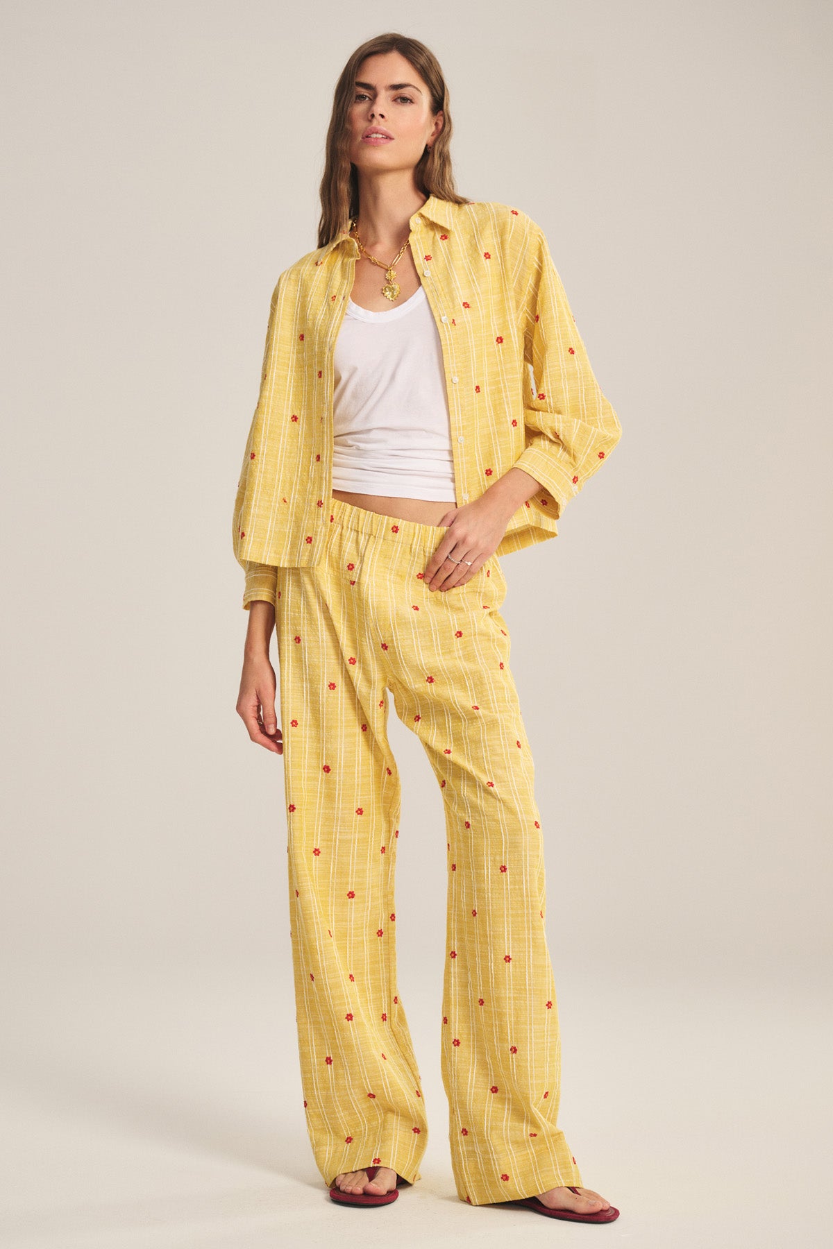 A woman stands against a plain background wearing Velvet by Graham & Spencer's MADELYN PANT with small red dots, paired with a loose yellow checkered shirt, white tank top, and sandals.