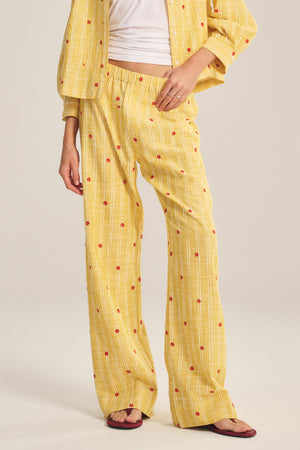 A person wears the MADELYN PANT by Velvet by Graham & Spencer—relaxed fit yellow pants with a matching long-sleeve shirt featuring red dots, white lines, a white top underneath, and sandals, posed against a plain background.