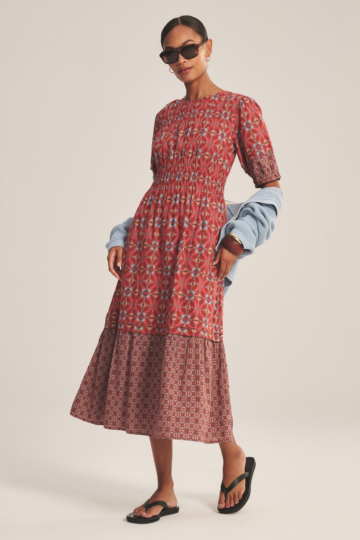 A woman stands against a plain background wearing the MAELA DRESS by Velvet by Graham & Spencer—a red patterned lightweight cotton dress with a tiered hem—paired with black sandals, sunglasses, and a light blue sweater draped over her arms.
