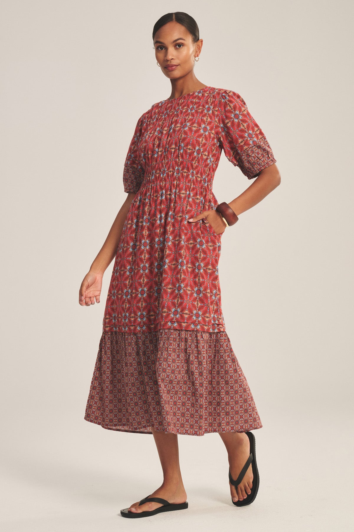 A woman wears the MAELA DRESS by Velvet by Graham & Spencer—a red patterned, lightweight cotton dress with puffed sleeves and a tiered skirt—paired with black sandals and a brown bangle.-41089019314369