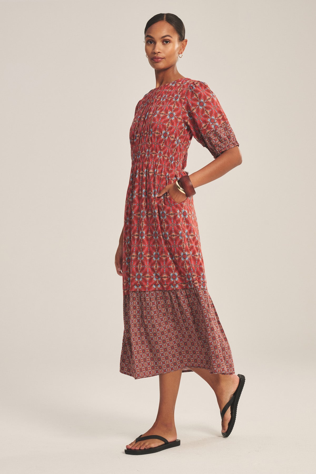 A person wearing the MAELA DRESS by Velvet by Graham & Spencer—a red patterned, short-sleeve, ankle-length dress with a smocked waist—paired with black sandals, stands sideways with one hand in a pocket.-41089019248833