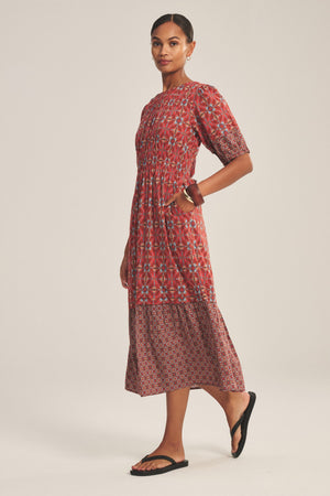A person wearing the MAELA DRESS by Velvet by Graham & Spencer—a red patterned, short-sleeve, ankle-length dress with a smocked waist—paired with black sandals, stands sideways with one hand in a pocket.