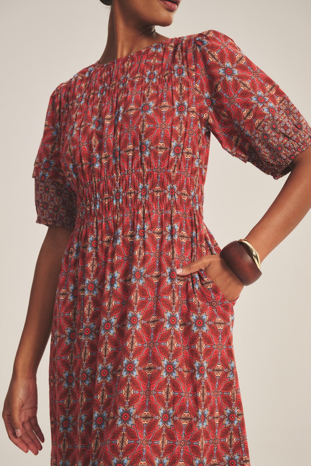 A woman wears the MAELA DRESS by Velvet by Graham & Spencer, a red lightweight cotton dress with blue and white geometric patterns, short puff sleeves, smocked waist, tiered hem, one hand in pocket, and a brown bangle.-41089019216065