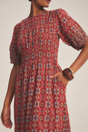 A woman wears the MAELA DRESS by Velvet by Graham & Spencer, a red lightweight cotton dress with blue and white geometric patterns, short puff sleeves, smocked waist, tiered hem, one hand in pocket, and a brown bangle.