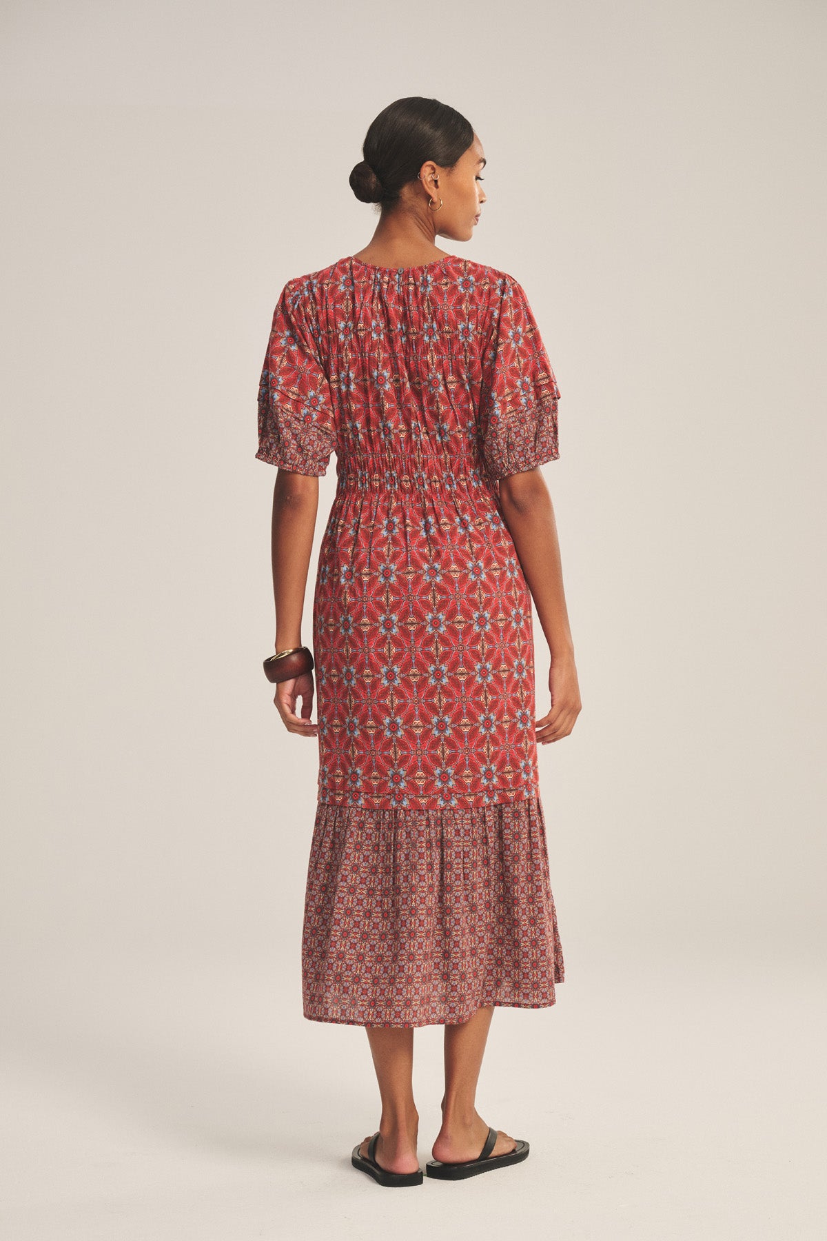 A woman faces away, wearing the MAELA DRESS by Velvet by Graham & Spencer—a red patterned, lightweight cotton dress with short sleeves and an elastic waist—paired with flat black sandals against a neutral background.-41089019183297
