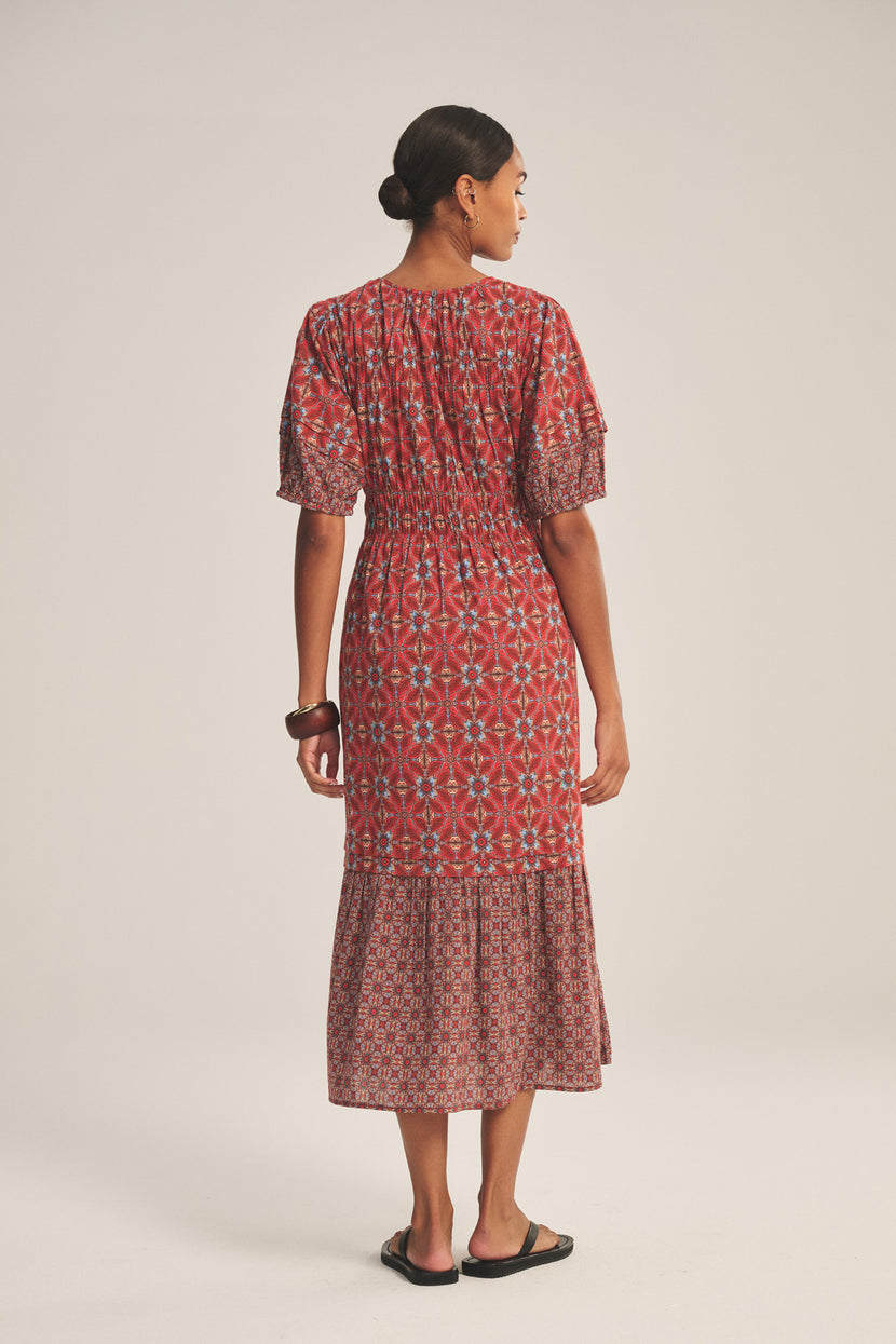 A woman faces away, wearing the MAELA DRESS by Velvet by Graham & Spencer—a red patterned, lightweight cotton dress with short sleeves and an elastic waist—paired with flat black sandals against a neutral background.