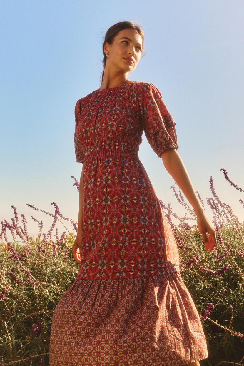 A woman wearing the MAELA DRESS by Velvet by Graham & Spencer stands in a field of purple flowers beneath a clear blue sky, gazing off to the side.