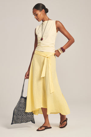 A woman wears the MALINA LINEN SKIRT by Velvet by Graham & Spencer, styled with a sleeveless top, patterned tote bag, layered necklaces, bangles, and sandals, standing against a plain background.