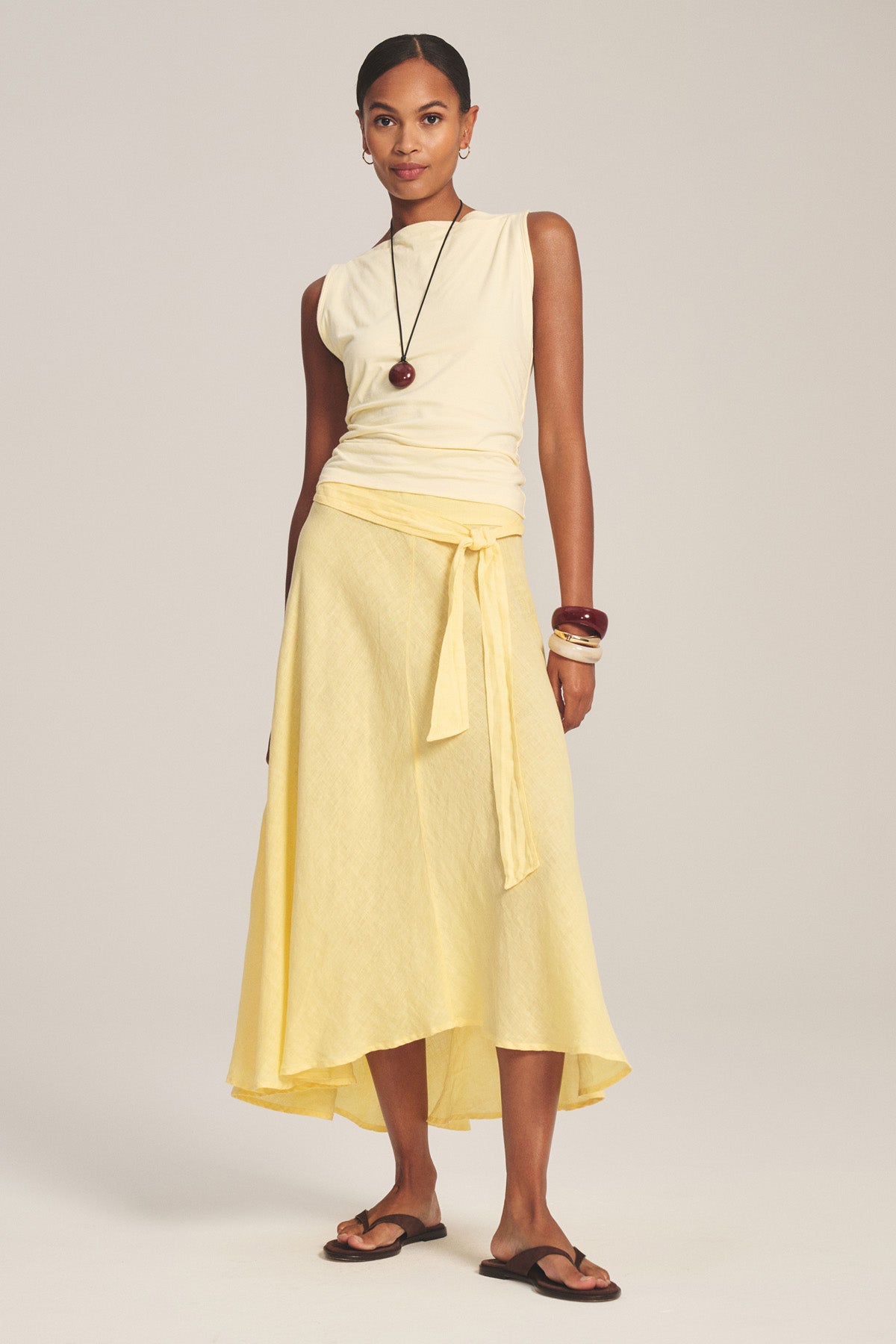 A woman stands against a plain background wearing the MALINA LINEN SKIRT by Velvet by Graham & Spencer—a yellow woven linen skirt with a versatile waist—styled with a sleeveless cream top, black sandals, bangles, and a long pendant necklace.-41128159379649