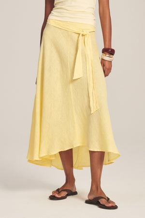 A person wears the MALINA LINEN SKIRT by Velvet by Graham & Spencer—a light yellow woven linen skirt with a waist tie—paired with a matching top, brown sandals, and stacked bangles, standing against a neutral background.