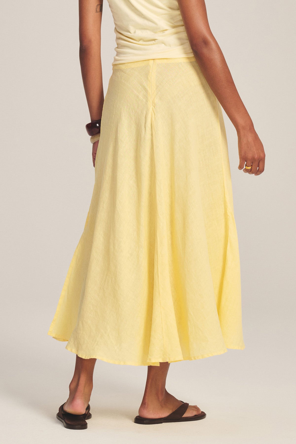 Model shown from the back against a plain background, wearing the MALINA LINEN SKIRT by Velvet by Graham & Spencer in yellow with a versatile waist fit, paired with a sleeveless top, black sandals, and bracelet.