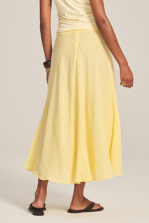 Model shown from the back against a plain background, wearing the MALINA LINEN SKIRT by Velvet by Graham & Spencer in yellow with a versatile waist fit, paired with a sleeveless top, black sandals, and bracelet.