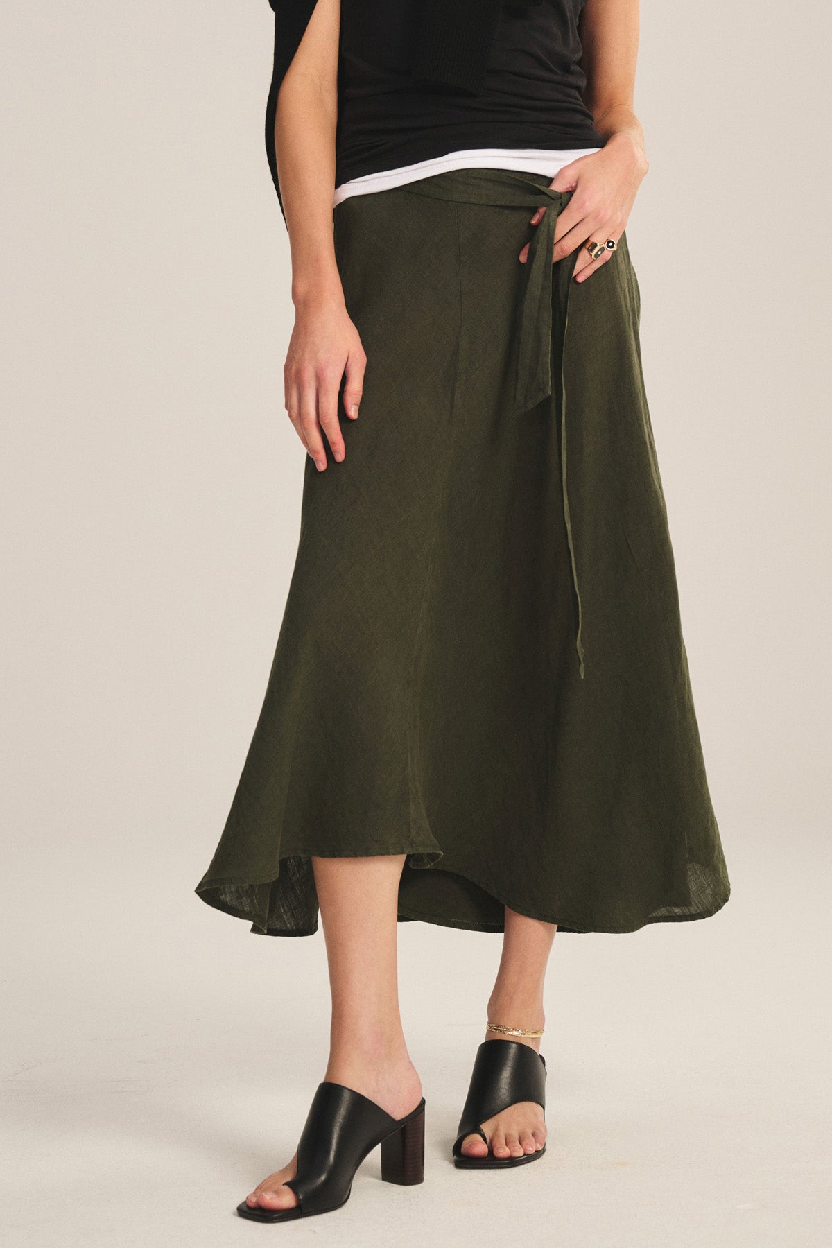Someone is wearing the MALINA LINEN SKIRT by Velvet by Graham & Spencer in olive green, paired with black open-toe heeled sandals, a black top, and a sweater draped over their shoulders.-40808415199425