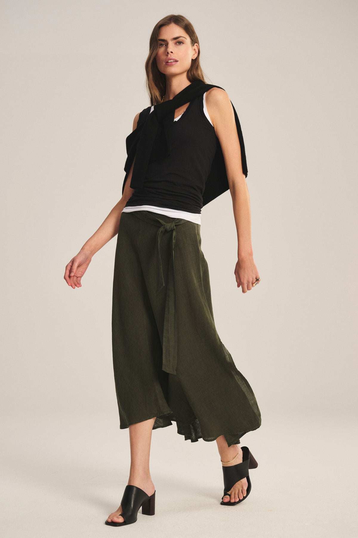 A person wearing a black sleeveless top, the MALINA LINEN SKIRT by Velvet by Graham & Spencer in olive green with a tie waist, and black heeled sandals poses against a plain background.-40808415166657