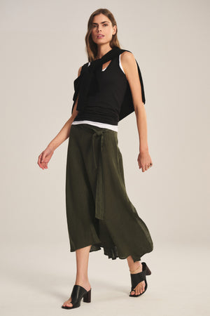 A person wearing a black sleeveless top, the MALINA LINEN SKIRT by Velvet by Graham & Spencer in olive green with a tie waist, and black heeled sandals poses against a plain background.