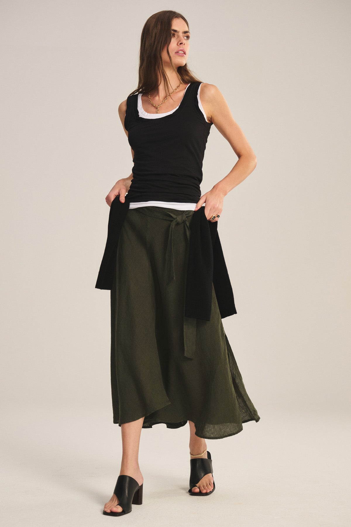 A woman wears the MALINA LINEN SKIRT by Velvet by Graham & Spencer—an olive green flowy skirt with a tie belt—paired with a black tank over a white shirt, black heeled sandals, and a black sweater draped at her waist.