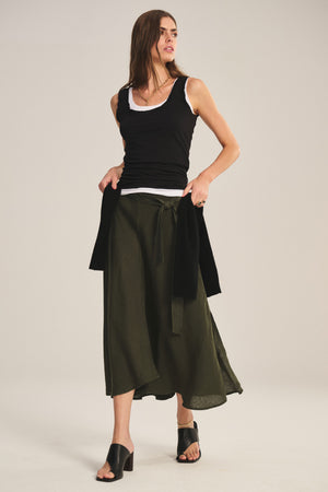 A woman wears the MALINA LINEN SKIRT by Velvet by Graham & Spencer—an olive green flowy skirt with a tie belt—paired with a black tank over a white shirt, black heeled sandals, and a black sweater draped at her waist.